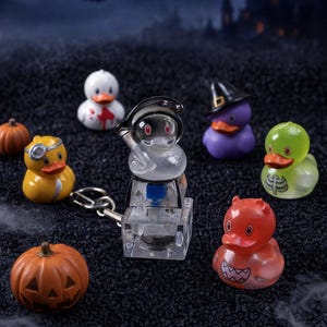 May include: A collection of small rubber ducks featuring various designs including a grim reaper, a witch, a skeleton, a doctor, and a red devil, alongside two miniature orange pumpkins and a clear mechanical keyboard switch keychain.