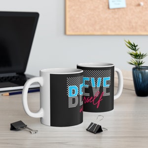 Może przedstawiać: Dwa białe ceramiczne kubki z czarno-białymi wzorami w kropki. Kubek po lewej stronie ma napis "Believe in yourself" niebieskimi i różowymi literami. Kubek po prawej stronie ma napis "Believe" niebieskimi literami.