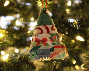 May include: A handmade Christmas tree ornament made of layered fabric. The ornament features a patchwork design with various patterns, including candy canes, bows, and geometric shapes. It hangs from a red string against a backdrop of a lit Christmas tree.