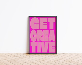 May include: A framed art print with the words "GET CREATIVE" in large, pink, bold letters against a bright pink background. The black frame is rectangular. The artwork is displayed against a white wall and a wooden floor.
