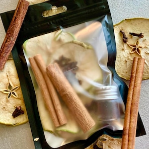 May include: A black pouch containing dried apple slices, cinnamon sticks, and a small jar. The pouch is surrounded by dried apple slices, star anise, and cloves. The cinnamon sticks are a rich brown color.