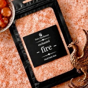 May include: A black resealable pouch containing light orange bath dust, labeled Fairy Apothecary enchanted fire ritual bath dust. The pouch is surrounded by loose orange salt crystals, a small bowl of orange stones, and a bronze phoenix figurine.