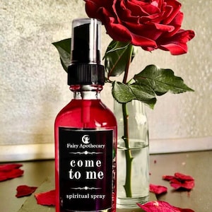 May include: A red glass bottle of "come to me" spiritual spray with a black label, next to a red rose in a clear vase. The bottle has a black cap and the label reads "Fairy Apothecary". Red rose petals are scattered around the base.