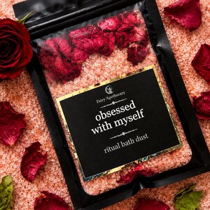 May include: A black pouch containing pink bath salts and dried red rose petals. The label features the text Fairy Apothecary, obsessed with myself, and ritual bath dust in white font against a black background with a gold border.
