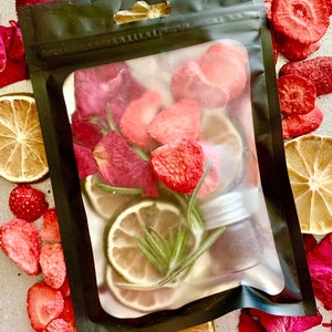 May include: A black pouch filled with dried strawberries, rose petals, and lime slices. A small glass bottle with a silver lid is visible inside the pouch. Dried strawberries, rose petals, and lemon slices surround the pouch on a light gray surface.