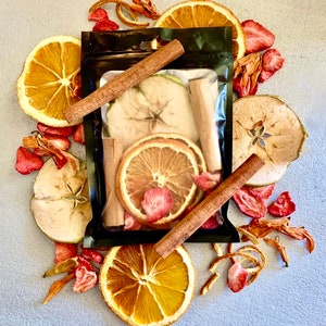 Op de afbeelding: Een zwarte zak gevuld met gedroogde appelschijfjes, sinaasappelschijfjes, aardbeien en kaneelstokjes. De zak is omgeven door extra gedroogd fruit en kruiden, wat een kleurrijke en aromatische presentatie creëert. De opstelling staat tegen een lichtgrijze achtergrond.