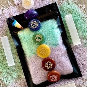 May include: A black pouch filled with light green and light purple bath salts, topped with five round stones in blue, green, yellow, red, and brown, each featuring a gold etched symbol. A multi-colored crystal pendulum and white selenite wands.