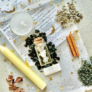 May include: A Pet Protection Spell Jar kit with a white candle, a yellow taper candle, a small glass bottle filled with green stones, and a bag of dried herbs. The kit includes a printed guide with instructions. The background is a white cloth with gold stars and crescent moons.