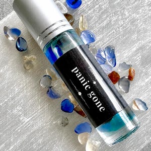 May include: A clear glass roller bottle with a silver cap and a black label featuring the white text "panic gone". The bottle contains blue liquid and small blue stones, surrounded by various small, polished, multi-colored decorative stones.