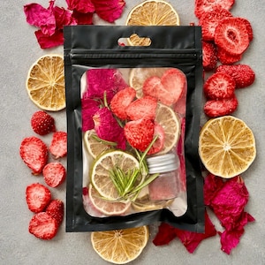 May include: A black resealable pouch containing a mix of dried strawberry slices, dehydrated lemon wheels, vibrant pink rose petals, fresh green rosemary sprigs, and a small glass jar filled with a fine pink powder, surrounded by loose dried fruit.