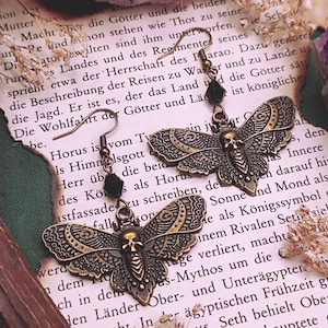 Könnte beinhalten: Antike, messingfarbene Ohrringe in Form von Motten mit filigranen Flügeldetails und einem Totenkopf-Design. Jeder Ohrring hat einen schwarzen Perlenakzent. Die Ohrringe liegen auf Vintage-Papier mit deutschem Text, umgeben von Trockenblumen.