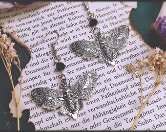 Puede incluir: Pendientes de plata con un diseño de calavera y mariposa. Los pendientes tienen cuentas negras y cuelgan de un libro con texto.