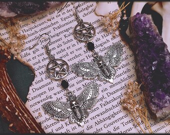 Puede incluir: Pendientes de plata con un amuleto de pentagrama y un diseño de mariposa detallado. Los pendientes tienen cuentas negras y cuelgan de un libro con texto sobre la mitología egipcia antigua.