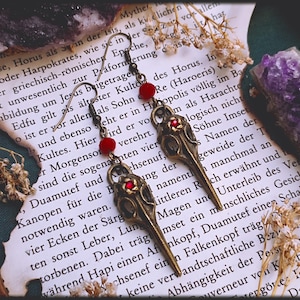 Könnte beinhalten: Ein Paar bronzefarbene Ohrringe mit einem Vogelschädel-Design und roten Glasperlen. Die Ohrringe hängen an einem Buch mit Text über die ägyptische Mythologie.