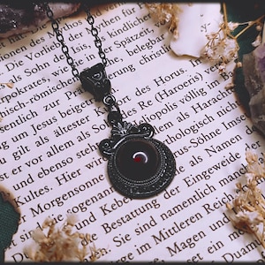 Puede incluir: Collar con colgante negro con un diseño circular adornado y un centro rojo oscuro. El collar está sobre una cadena y descansa sobre una página de texto con flores secas y un cristal púrpura.