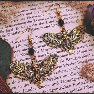 Könnte beinhalten: Goldfarbene Ohrringe in Form von Motten mit detaillierten Verzierungen und schwarzen Perlen. Sie liegen auf einem Hintergrund aus altem Papier mit Text, was ein gotisches oder mystisches Thema erzeugt.