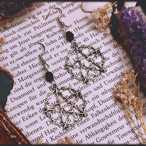 Könnte beinhalten: Silberne Pentagramm-Ohrringe mit schwarzen Perlen, präsentiert auf einer Vintage-Buchseite mit getrockneten Blumen und einem lila Kristall. Die Ohrringe haben ein gotisches Design.