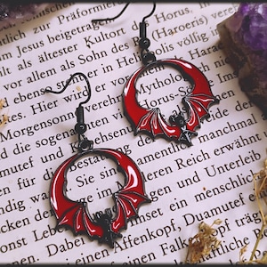 Könnte beinhalten: Ein Paar rote und schwarze Fledermaus-Ohrringe. Die Ohrringe sind in Form eines Halbmondes mit Fledermausflügeln geformt. Die Fledermausflügel sind rot mit schwarzen Umrissen. Die Ohrringe hängen an schwarzen Haken.