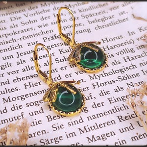 May include: Gold-coloured earrings with a dragonfly design, featuring a round, deep green stone. The earrings are set against a backdrop of aged paper with text, and dried flowers.