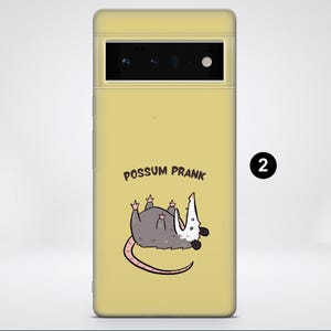 Puede incluir: Una funda de tel&eacute;fono amarilla con un opossum de dibujos animados en ella. El opossum est&aacute; tumbado de espaldas con las patas en el aire. El texto "Possum Prank" est&aacute; impreso en la funda.