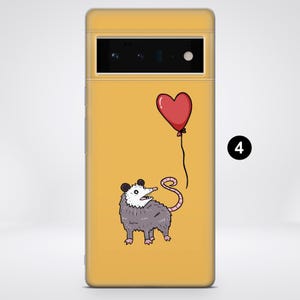 Puede incluir: Una funda de tel&eacute;fono amarilla con una ilustraci&oacute;n de dibujos animados de una zarig&uuml;eya sosteniendo un globo rojo en forma de coraz&oacute;n. La zarig&uuml;eya tiene una cara blanca y un cuerpo gris.