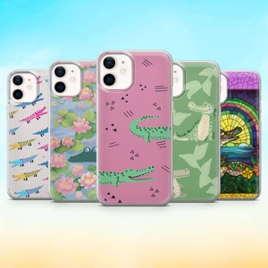 Op de afbeelding: Vijf verschillende telefoonhoesjes met alligator-ontwerpen. De hoesjes zijn transparant en laten de camera van de telefoon zien. De ontwerpen omvatten een roze hoesje met zwarte geometrische vormen en alligators, een groen hoesje met een jungle-scène en alligators, een blauw hoesje met een bloemenpatroon en alligators, een wit hoesje met een kleurrijk alligator-patroon en een hoesje met een glas-in-loodontwerp van een alligator.
