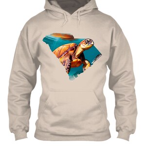 May include: A tan hoodie featuring a colorful sea turtle design within the outline of South Carolina. The turtle is depicted in shades of brown, orange, and yellow against a blue and teal background. The text "SOUTH CAROLINA" is visible.