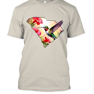 May include: A beige t-shirt with a graphic print of a hummingbird flying over a floral design inside the outline of the state of South Carolina.