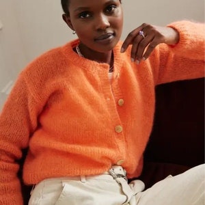 May include: A bright orange fuzzy cardigan with a button closure. The cardigan is worn over a white shirt and white pants.