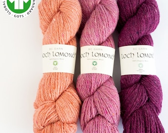 May include: Three balls of organic wool yarn in shades of peach, pink, and deep purple. Each ball has a white label with the text "BC Garn Loch Lomond 100% Organic Wool". The GOTS organic textile standard logo is visible.