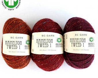 May include: Three skeins of yarn in different colors: rust, burgundy, and dark red. Each skein has a label that reads "BC Garn Hamilton Tweed 1 90% Wool / 10% Viscose" and a green GOTS logo. The label also states that the yarn is made with 90% organic materials certified by BCS.