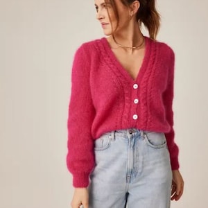 Könnte beinhalten: Ein rosa flauschiger Cardigan mit V-Ausschnitt und Knopfleiste. Der Cardigan hat ein Zopfmuster und wird über einer hellblauen Jeans getragen.