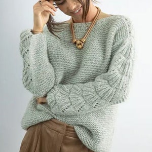 Peut inclure: Un pull en tricot vert clair avec un motif de tricot torsadé épais sur les manches. Le pull a un col rond et est porté avec une jupe marron.