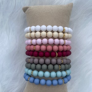 Peut inclure: Une pile de huit bracelets avec des perles de différentes couleurs. Les bracelets sont faits de perles en bois avec une finition mate. Chaque bracelet a un accent doré.