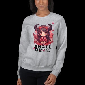 Op de afbeelding: Een grijs sweatshirt met een grafische print van een cartoonmeisje met hoorns en rode ogen. Het meisje zit op een troon gemaakt van schedels en is omringd door andere schedels. De tekst "SMALL DEVIL" is onder de afbeelding gedrukt.
