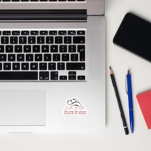 May include: A white laptop computer with a black keyboard and a sticker that says "Beth Dutton STATE OF MIND" with a drawing of a cowboy hat.