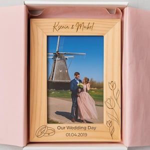 Puede incluir: Marco de fotos de madera con una foto de una pareja frente a un molino. El marco está grabado con los nombres "Ksenia & Maikel", el texto "Our Wedding Day" y la fecha "01.04.2019". El marco tiene diseños de tulipanes grabados.