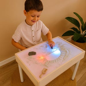 Può includere: Un bambino gioca con un tavolo di sabbia illuminato. Il tavolo ha una cornice in legno e gambe bianche. La sabbia è illuminata con luci colorate. Un'auto giocattolo e una paletta di legno sono sul tavolo.