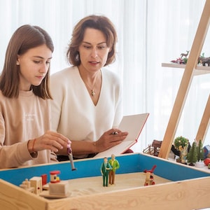 Puede incluir: Dos mujeres están sentadas a una mesa con una bandeja de arena. La bandeja contiene figuras y estructuras en miniatura. Una estantería de madera en el fondo muestra más artículos en miniatura. Las mujeres miran la bandeja y un cuaderno.