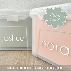May include: Two clear plastic storage boxes with personalized name decals. The box on the left has a green background and the name "Joshua" in white lettering. The box on the right has a peach background and the name "Nora" in white lettering. The box on the right also has a green cloud-shaped decal with the text "name decal" in white lettering.