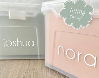 Op de afbeelding: Twee doorzichtige plastic opbergdozen met gepersonaliseerde naamlabels. De doos aan de linkerkant heeft een groene achtergrond en de naam "Joshua" in witte letters. De doos aan de rechterkant heeft een perzikkleurige achtergrond en de naam "Nora" in witte letters. De doos aan de rechterkant heeft ook een groen wolkvormig label met de tekst "name decal" in witte letters.