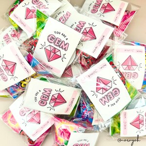 Puede incluir: Una pila de peque&ntilde;as bolsas de pl&aacute;stico transparente llenas de caramelos y peque&ntilde;as tarjetas rosas y blancas con el texto "You are a GEM" y un gr&aacute;fico de diamante rosa. Las tarjetas est&aacute;n firmadas "From: Joshua".
