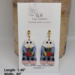 May include: A pair of dangle earrings featuring a white ghost figure wearing a blue and black plaid dress and holding a small Christmas tree. The earrings are 1.40 inches long and .80 inches wide.