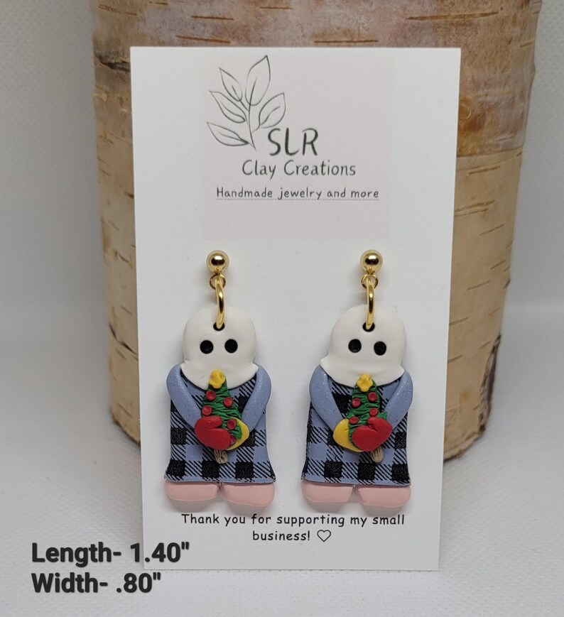 May include: A pair of dangle earrings featuring a white ghost figure wearing a blue and black plaid dress and holding a small Christmas tree. The earrings are 1.40 inches long and .80 inches wide.