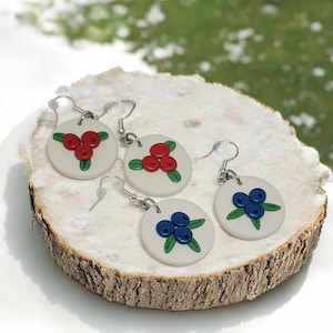Peut inclure: Quatre paires de boucles d'oreilles artisanales sur le thème des fruits. Chaque boucle d'oreille présente une base circulaire blanche avec un groupe de baies rouges ou de myrtilles bleues, ainsi que des feuilles vertes. Les boucles d'oreilles sont exposées sur une tranche de bois.