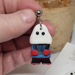 May include: A pair of polymer clay earrings featuring a white figure with black eyes, red mittens, and a blue jacket playing an accordion. The figure is wearing black pants and red shoes.