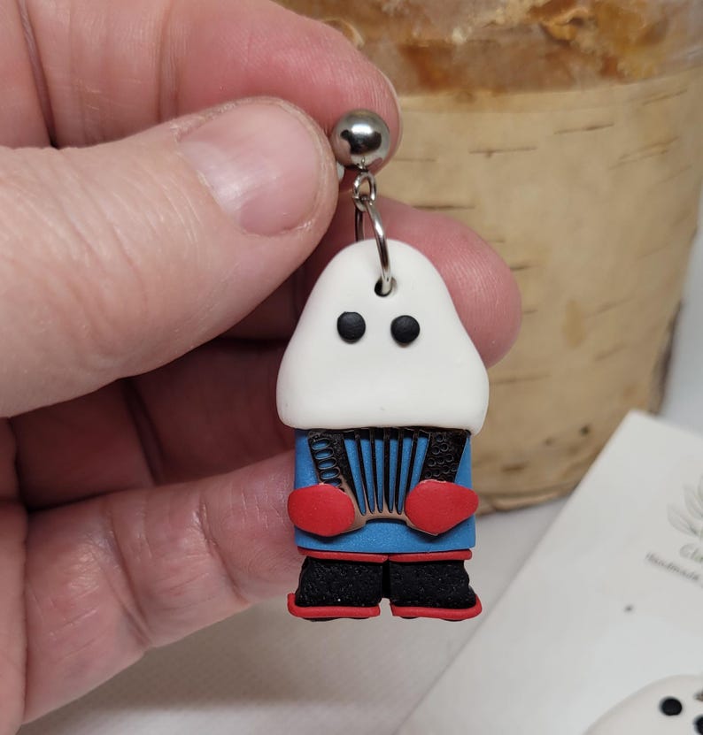 May include: A pair of polymer clay earrings featuring a white figure with black eyes, red mittens, and a blue jacket playing an accordion. The figure is wearing black pants and red shoes.