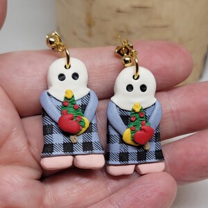 May include: A pair of dangle earrings featuring a ghost holding a small Christmas tree with red ornaments. The ghost is wearing a blue and black plaid dress. The earrings are made of polymer clay and have gold hooks.