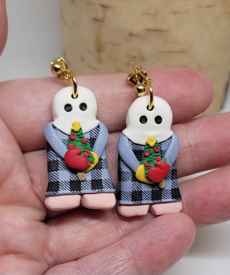 May include: A pair of dangle earrings featuring a ghost holding a small Christmas tree with red ornaments. The ghost is wearing a blue and black plaid dress. The earrings are made of polymer clay and have gold hooks.
