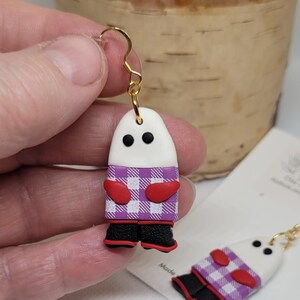 May include: A pair of white ghost-shaped earrings with purple and white checkered shirts, red mittens, and black boots. The earrings are made of polymer clay and have gold hooks.